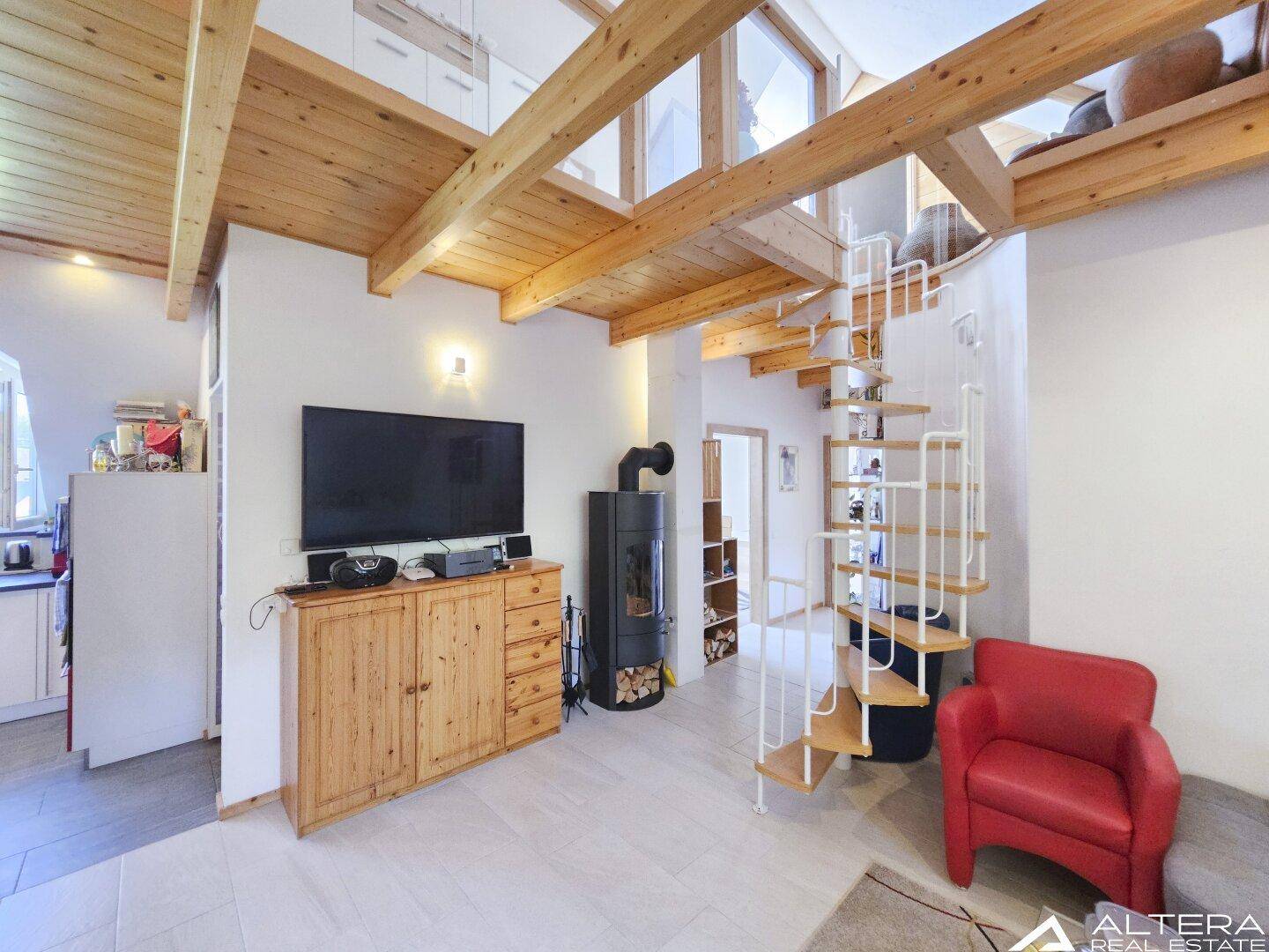 Wohnzimmer mit Kaminofen, TV-Möbel und Wendeltreppe, unter Holzbalkendecke.