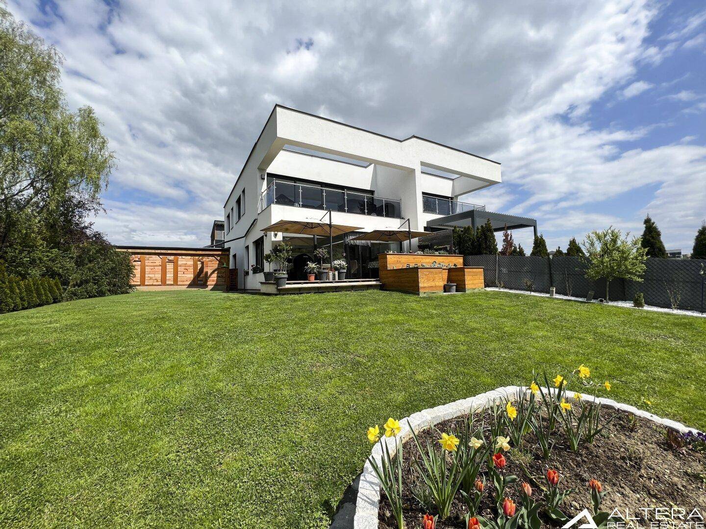 Moderne weiße Villa mit großem gepflegten Rasen und Blumenbeeten unter bewölktem Himmel.