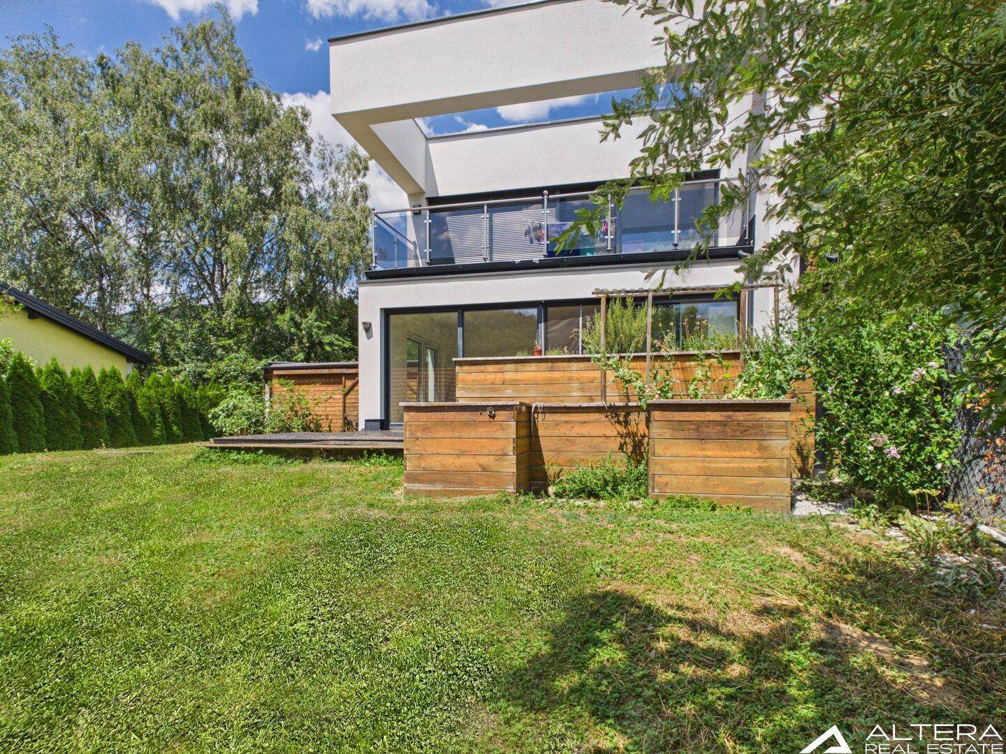 Seitlicher Blick auf die moderne Villa mit erhöhten Holzbeeten und weitläufigem grünen Gartenbereich.