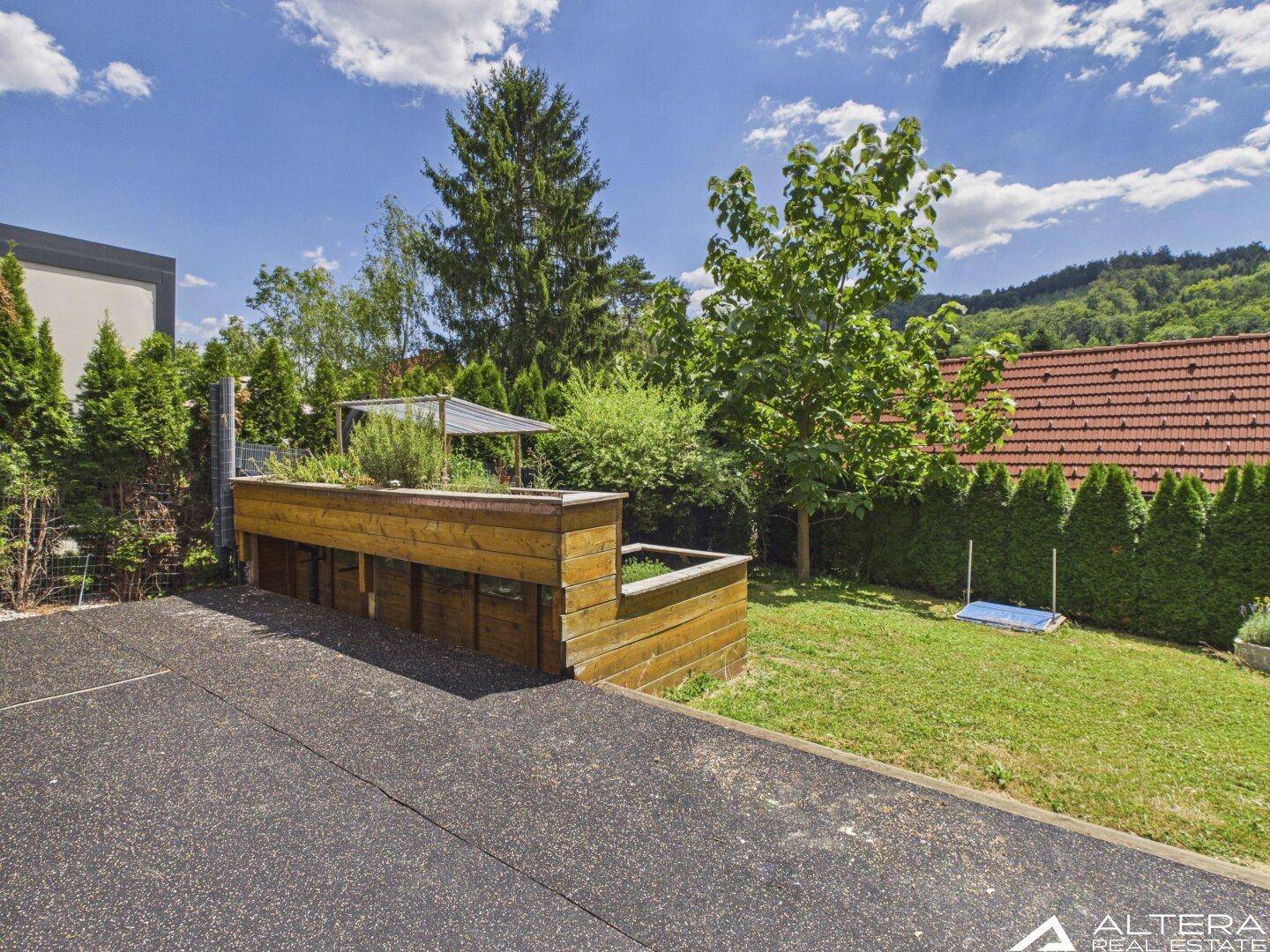 Hinterhofbereich mit gepflastertem Weg, erhöhten Holzbeeten und einem Trampolin, umgeben von Grün.