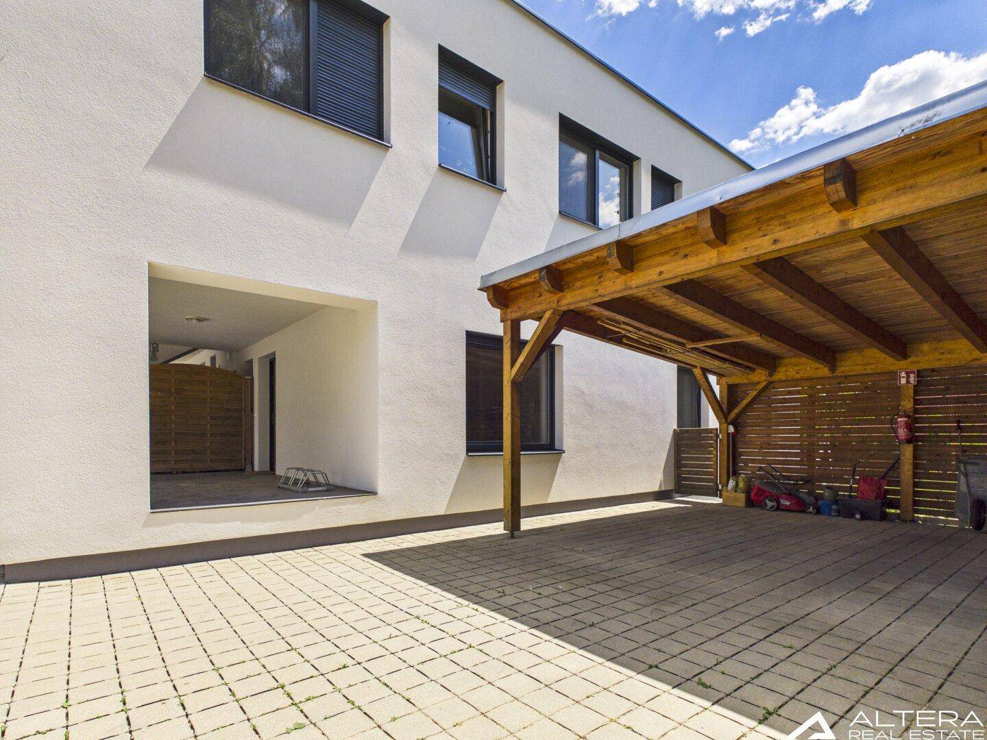 Moderne weiße Hausfassade mit Fenstern und einem hölzernen Carport auf gepflastertem Vorplatz.