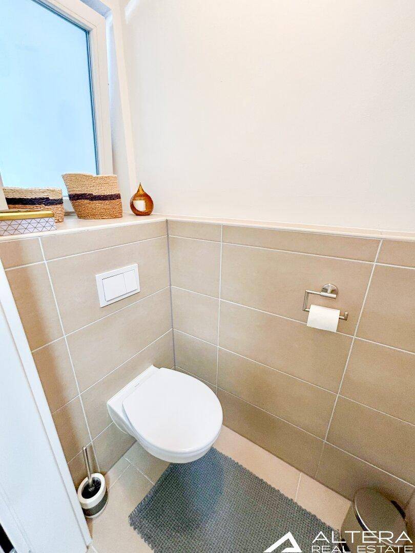 Kleines Gäste-WC mit beigefarbenen Fliesen, wandhängendem WC und einem Fenster für Tageslicht.