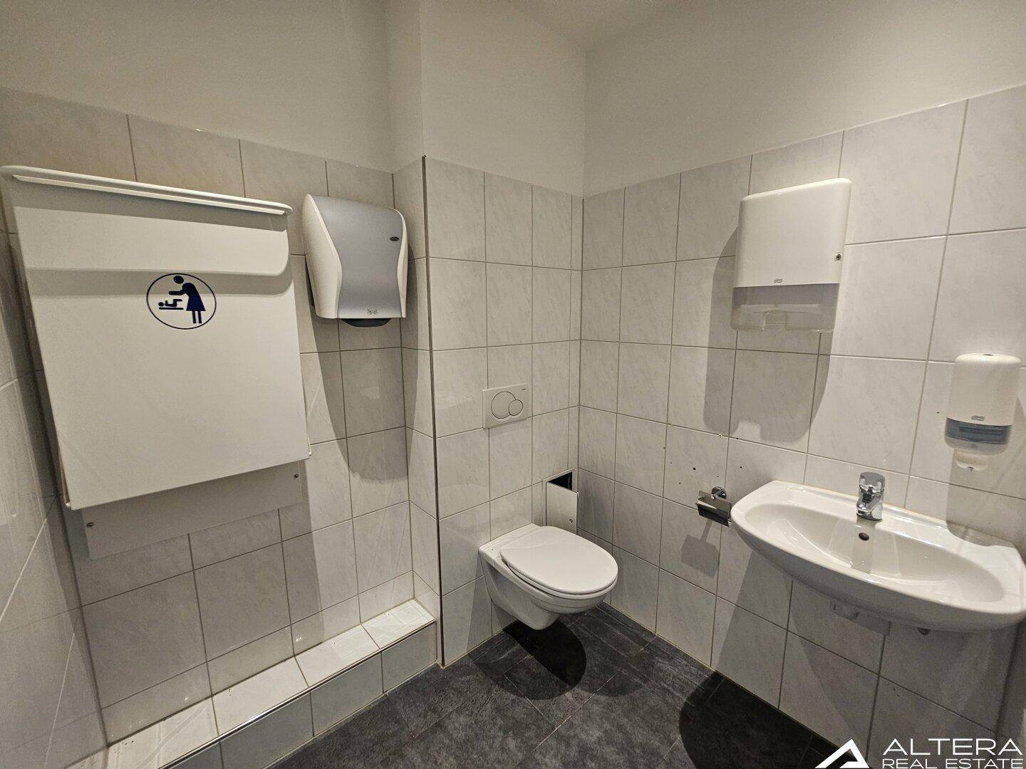 Saubere Toilettenkabine mit wandhängendem WC, kleinem Waschbecken und Wickeltisch für Babys.