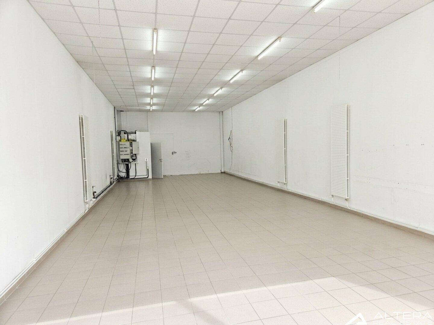 Geräumiger Gewerberaum mit Fliesenboden, Heizkörpern und sichtbaren Installationen an der Wand.