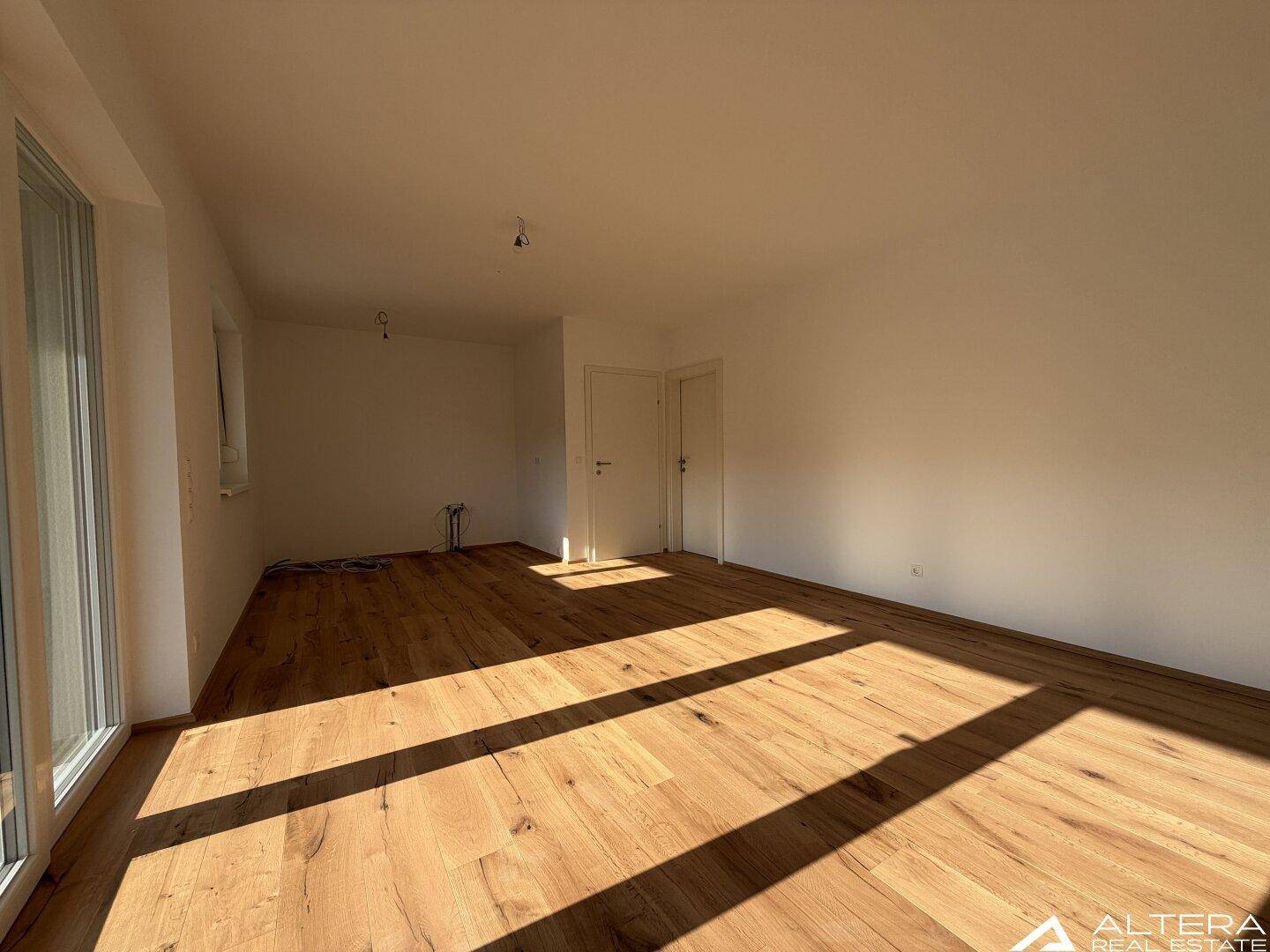 Geräumiges, leeres Wohnzimmer mit Holzboden und viel natürlichem Licht durch die Fenster.