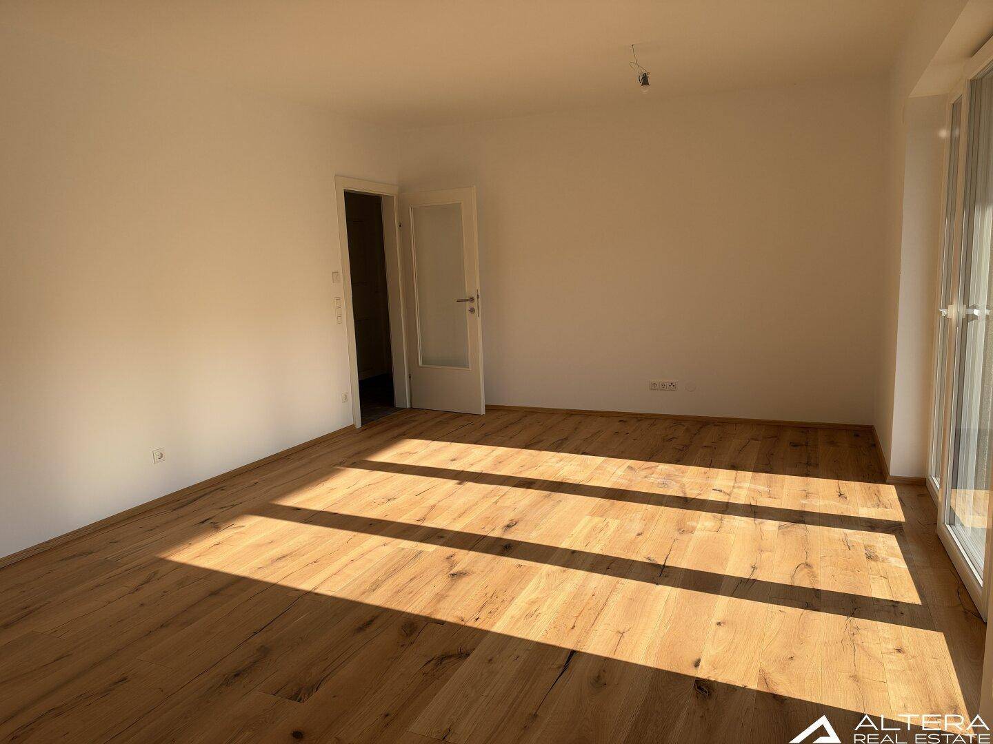 Leeres Wohnzimmer mit Holzboden und einer Tür, die zu einem weiteren Raum führt.