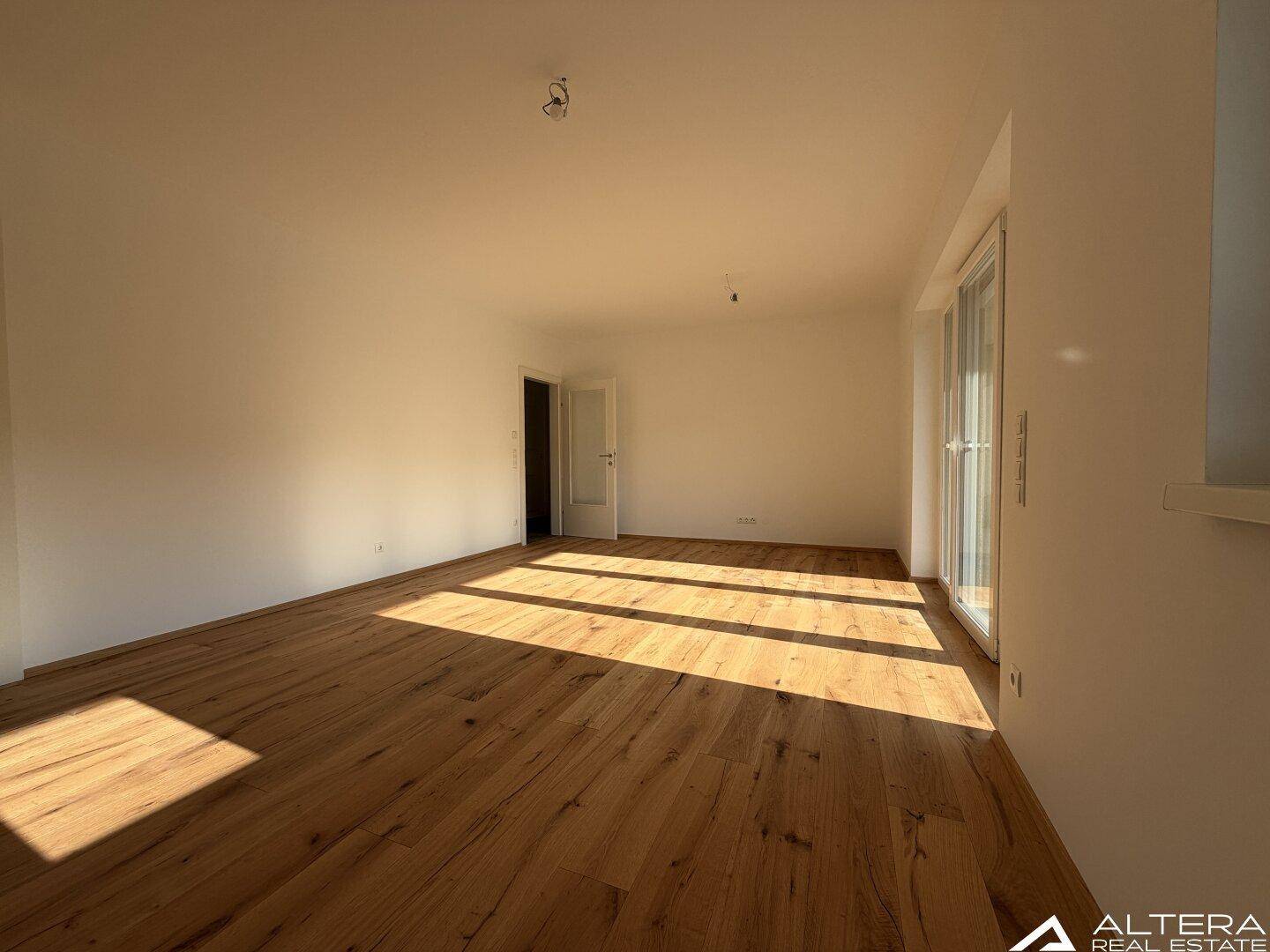 Heller Wohnbereich mit Holzboden und großen Fenstern, die viel Sonnenlicht hereinlassen.