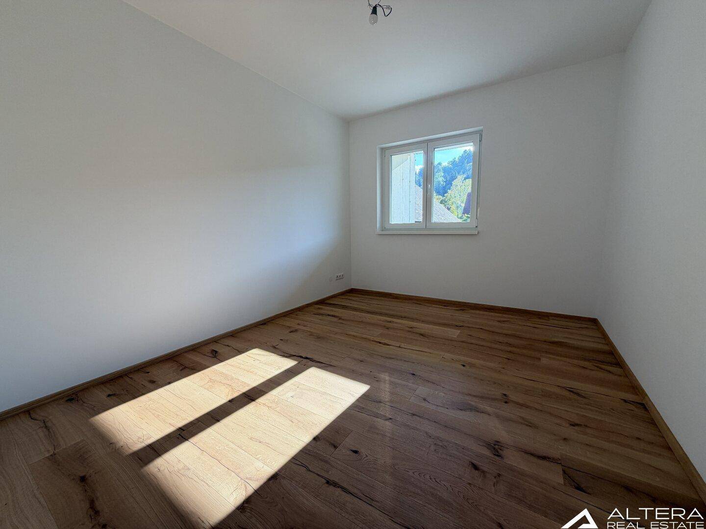 Helles Schlafzimmer mit Holzboden und Fenster, das einen Blick ins Grüne bietet.