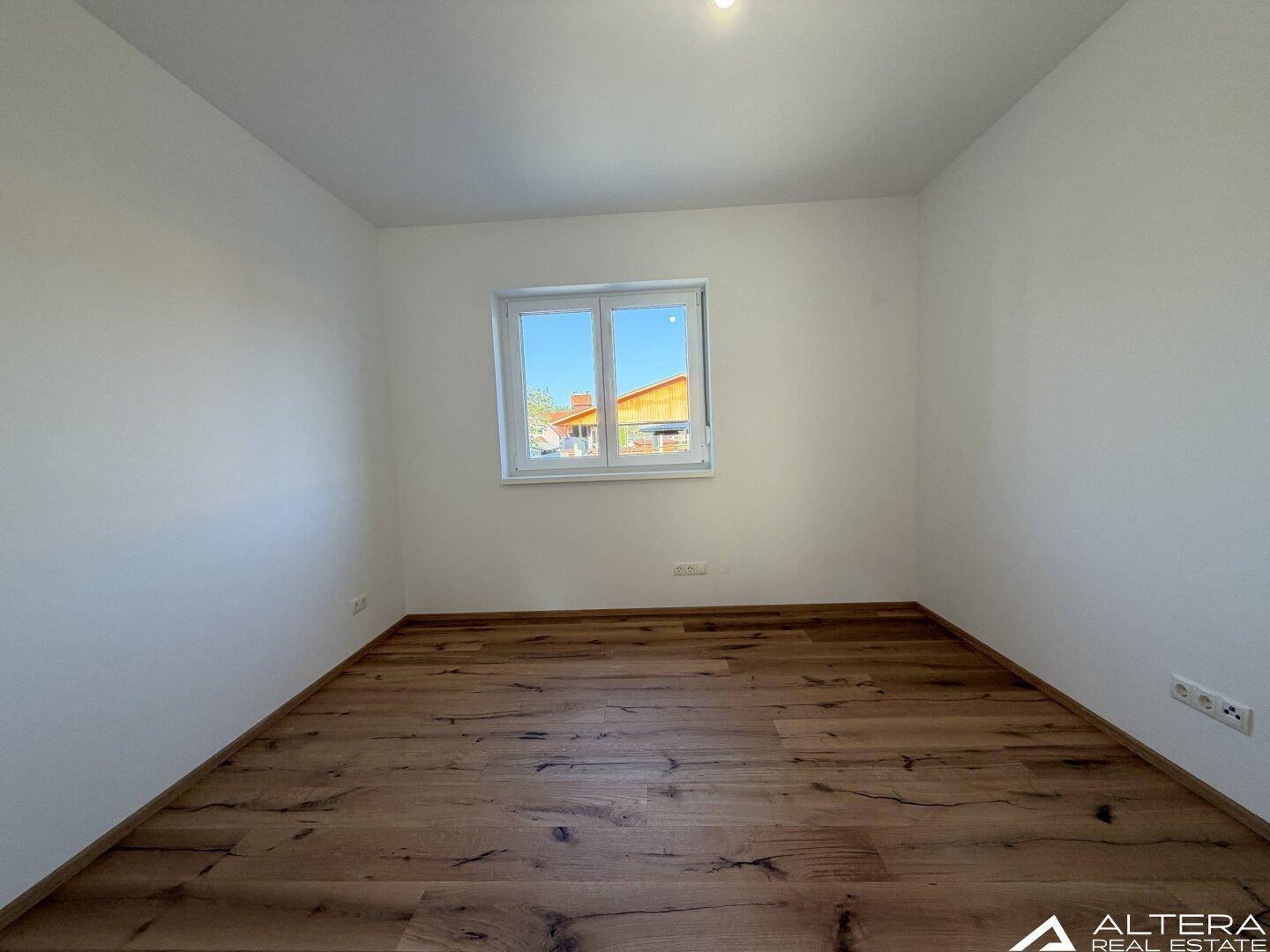 Leeres Schlafzimmer mit Holzboden und einem Fenster, das viel Tageslicht spendet.