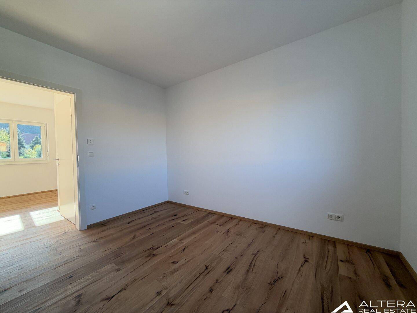 Schlafzimmer mit Holzboden und offener Tür, die den Blick in einen weiteren Raum freigibt.