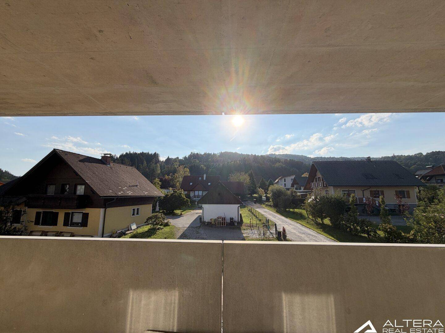 Sonniger Balkon mit Betonboden und Panoramablick auf die umliegende Natur und Häuser.