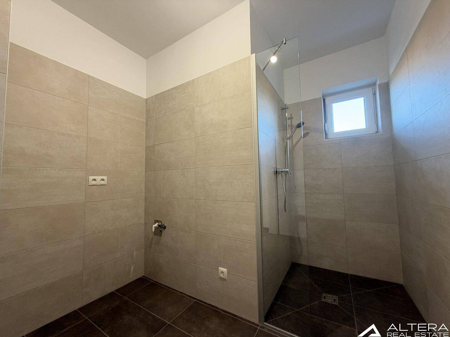 Badezimmer mit begehbarer Dusche, hellen Wandfliesen und dunklem Boden.