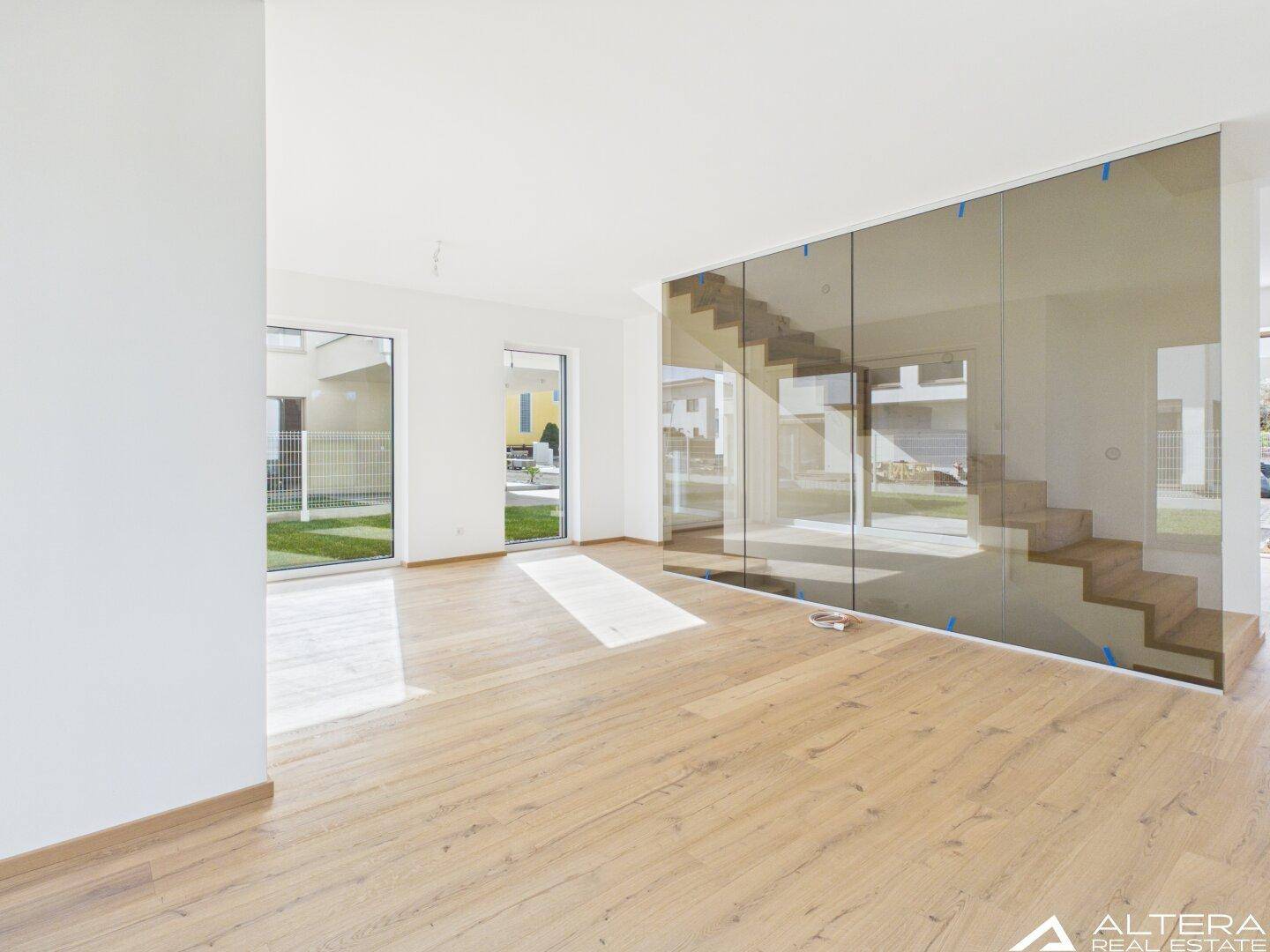Geräumiges Wohnzimmer mit Holzboden und moderner Glaswand, die eine Treppe umschließt.