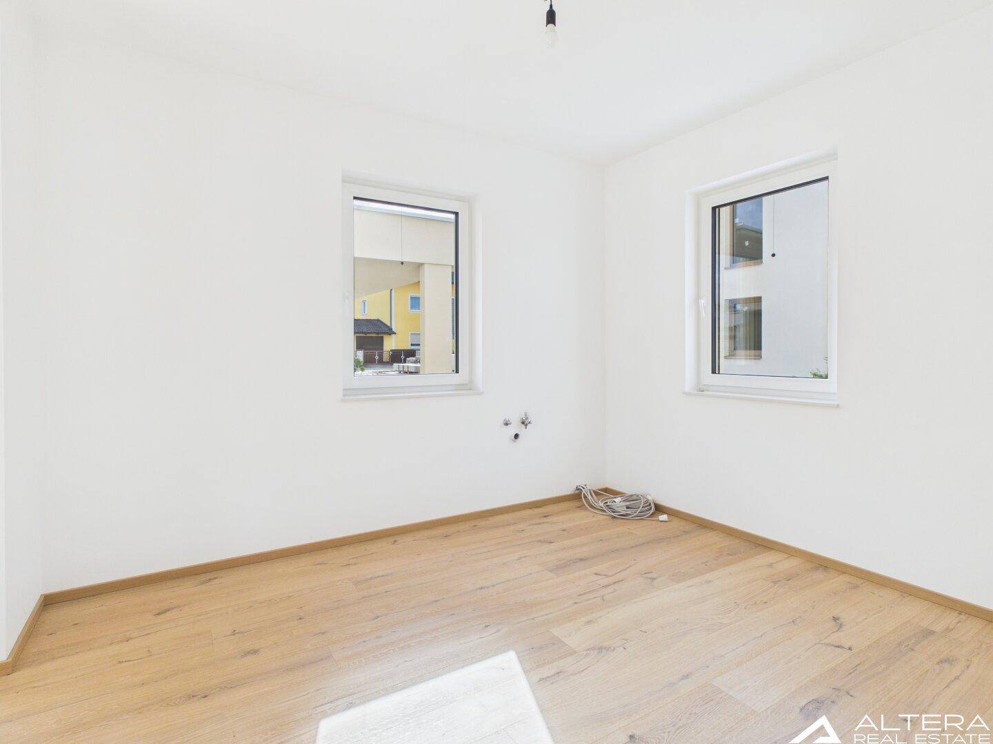 Geräumiges Zimmer mit Holzboden und viel Tageslicht durch zwei Fenster.