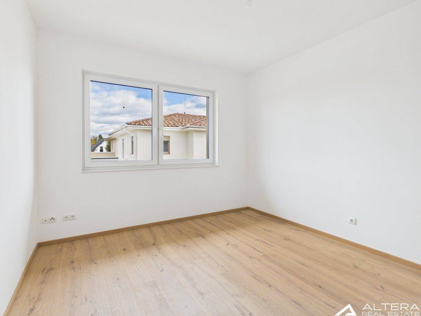 Helles Zimmer mit Holzboden und Fenster, das einen Blick auf die Nachbarschaft bietet.