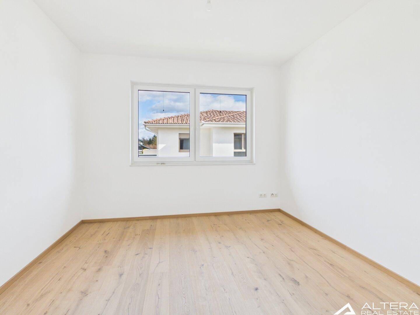 Geräumiges Zimmer mit Holzboden und großem Fenster, das viel Tageslicht spendet.