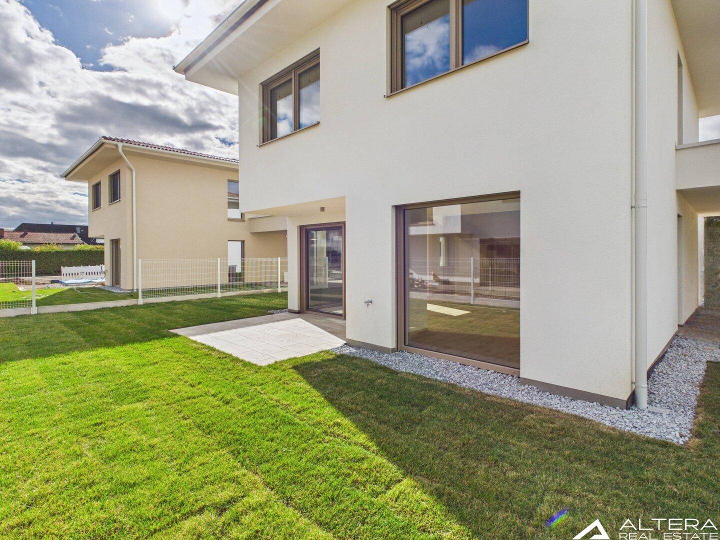Gartenbereich mit Rasenfläche und kleiner Terrasse, zugänglich durch große Fenster.