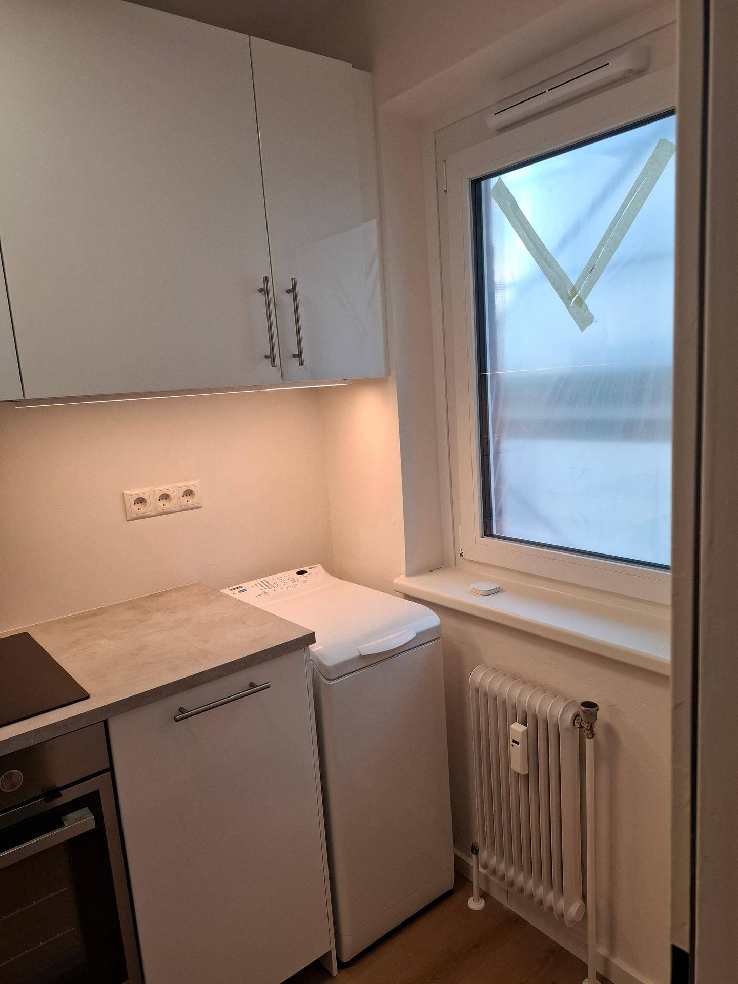 Kompakte Küche mit Fenster, Heizkörper und hellen Schränken, ideal für kleine Haushalte.