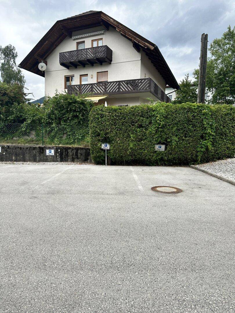 Wohnhaus mit mehreren Balkonen hinter einer hohen Hecke und einem Parkplatz davor.