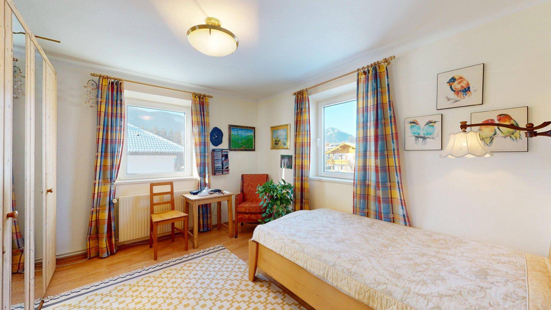 Helles Schlafzimmer mit Einzelbett, bunten Vorhängen und Bergblick durch die Fenster.
