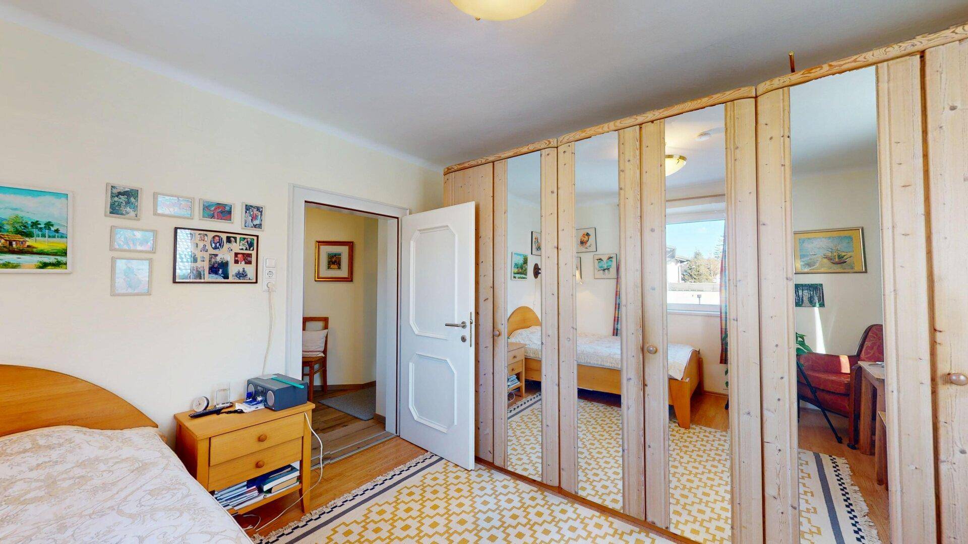 Schlafzimmer mit Spiegelschränken, Holzboden und Zugang zu einem weiteren Raum.