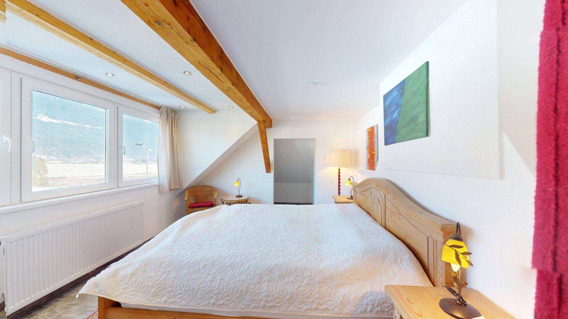 Schlafzimmer im Dachgeschoss mit sichtbaren Holzbalken, großen Fenstern und Bergblick.