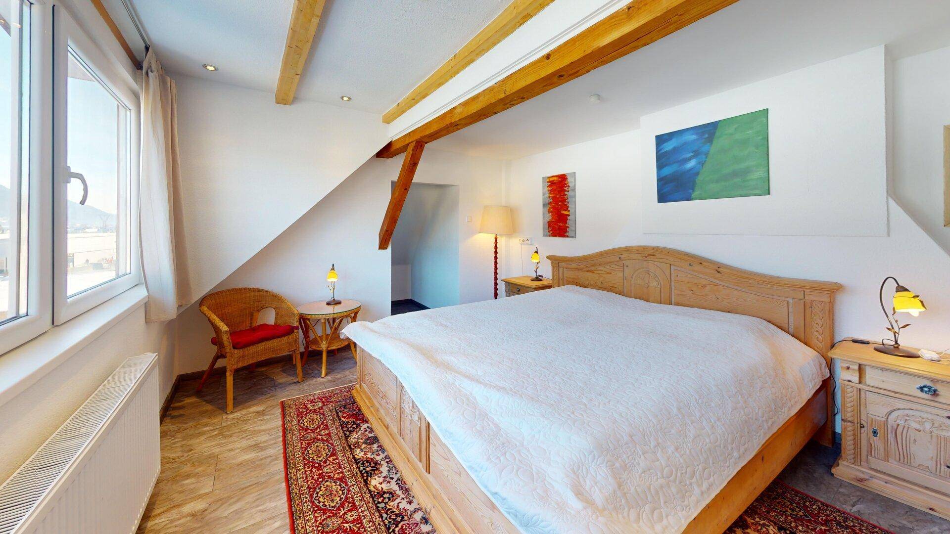 Helles Dachgeschoss-Schlafzimmer mit Holzbalken, Holzbett und Teppichboden.