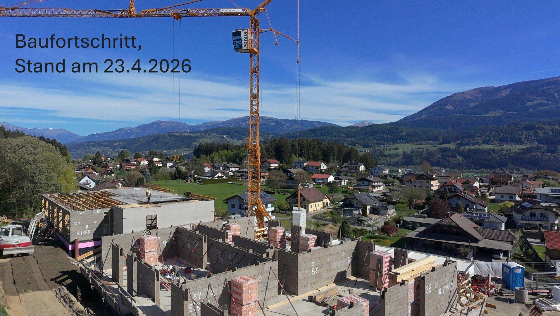 Luftaufnahme einer Baustelle mit Kran und im Bau befindlichen Gebäuden vor einer malerischen Berglandschaft.