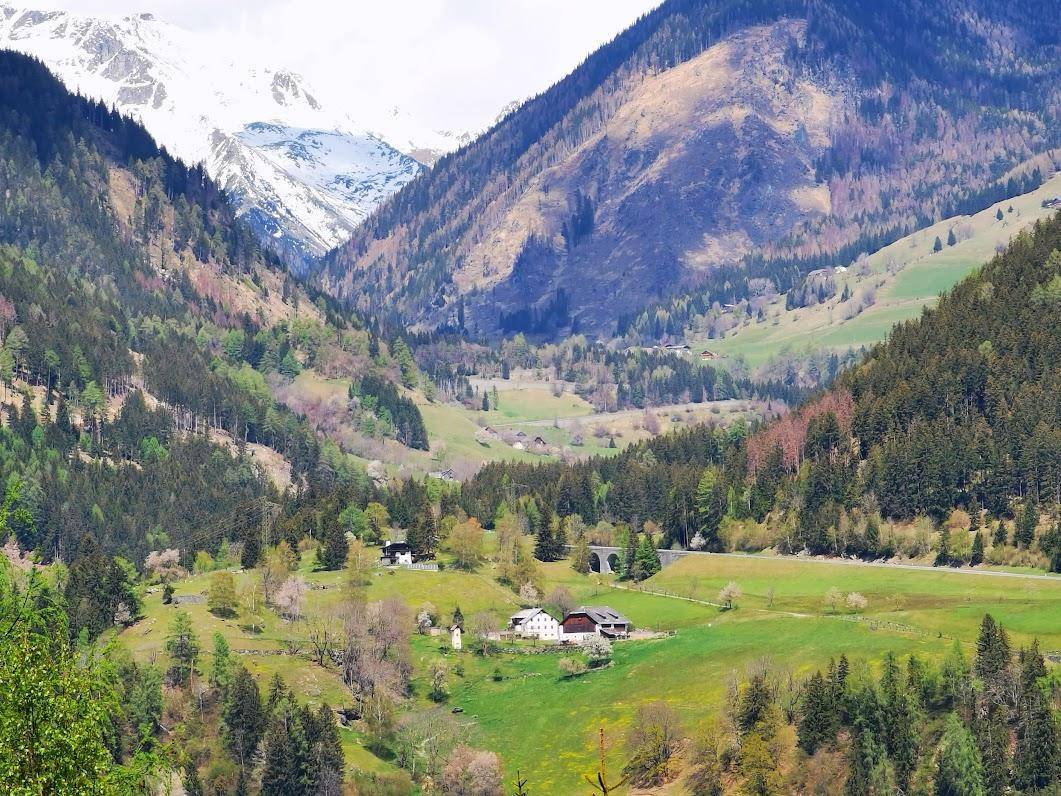Malerisches Tal mit verstreuten Häusern, grünen Wiesen und schneebedeckten Berggipfeln unter blauem Himmel.