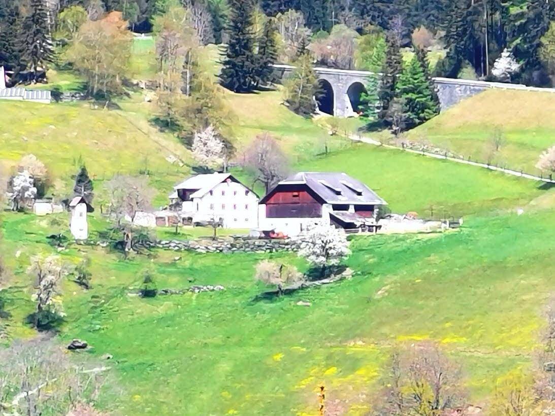 Gruppe von traditionellen Häusern in einer grünen Hügellandschaft mit einem historischen Viadukt im Hintergrund.