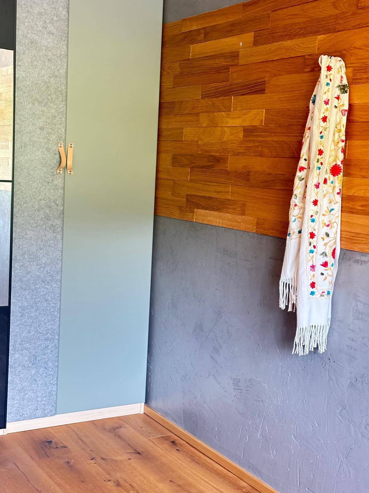 Detailansicht einer Wand mit Holzpaneelen, grauer Textur und einem farbenfrohen Schal als Dekoration.