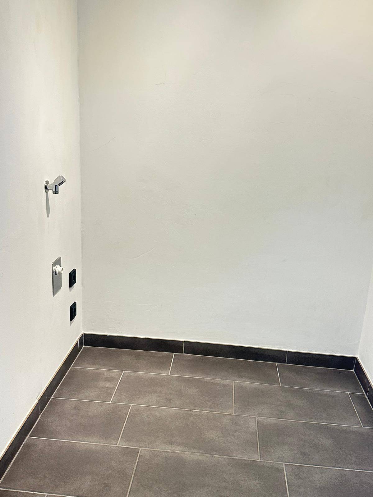 Leerer Raum mit Fliesenboden und Anschlüssen, ideal als Abstell- oder Hauswirtschaftsraum.