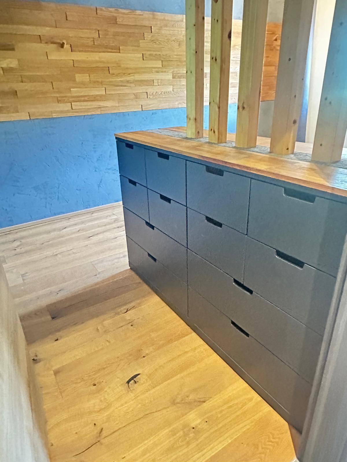 Detailaufnahme einer dunklen Kommode mit Holzplatte neben einer Holz-Raumtrennung auf hellem Holzboden.