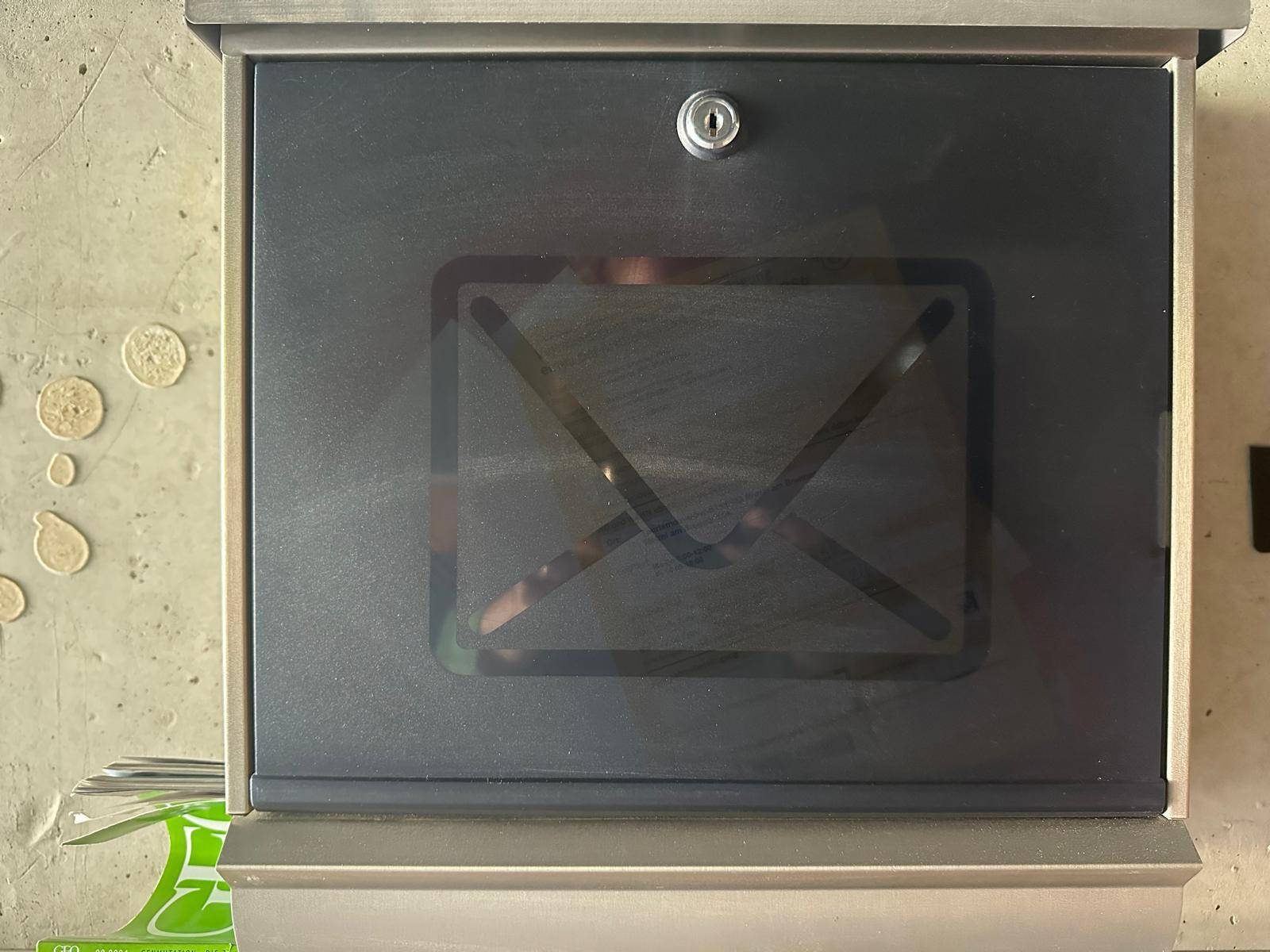 Moderner Briefkasten aus Metall mit stilisiertem Briefsymbol, an einer Betonwand montiert.