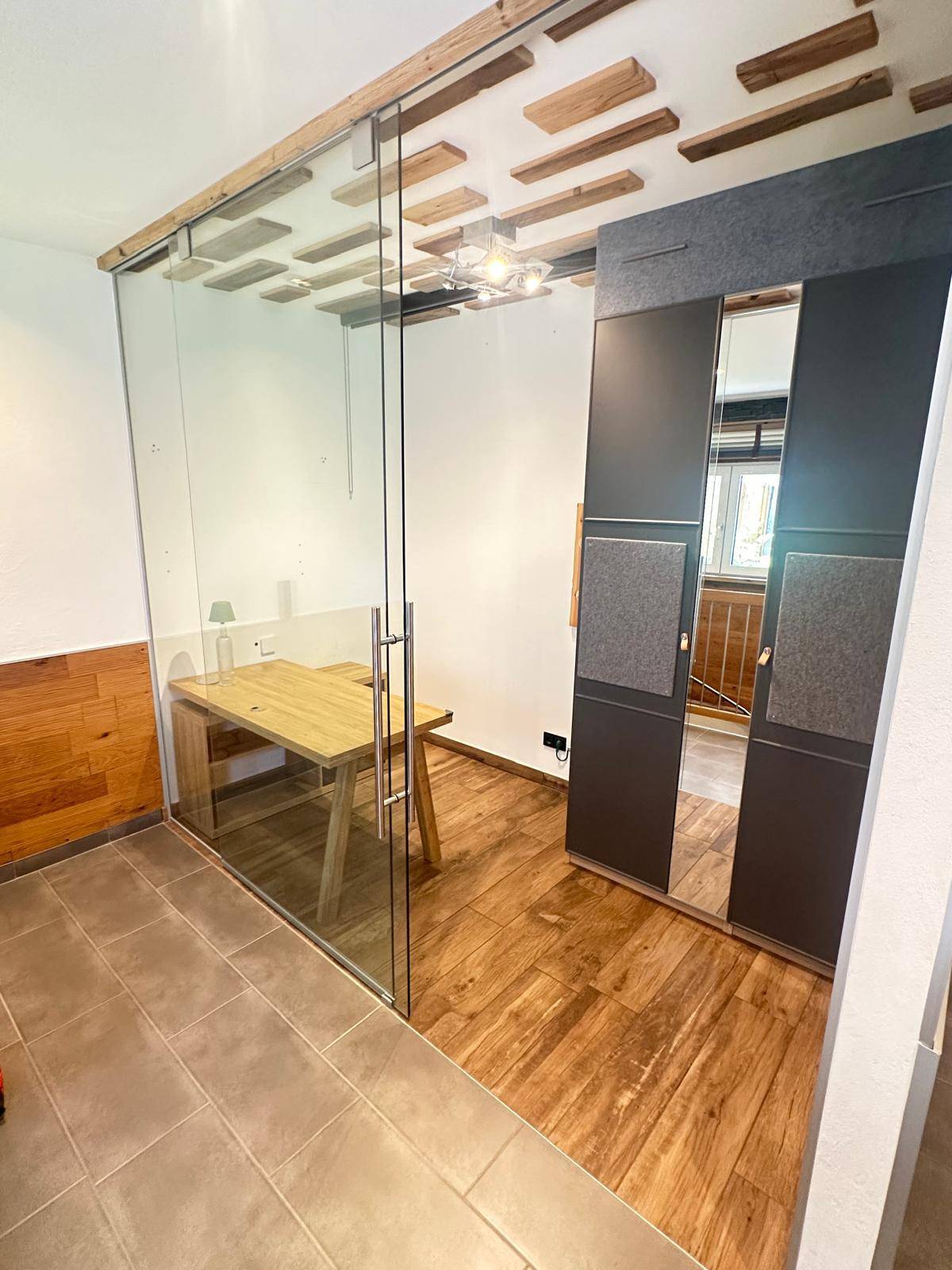 Modernes Büro mit Glastrennwand, Holztisch und unterschiedlichen Bodenbelägen, die den Bereich definieren.