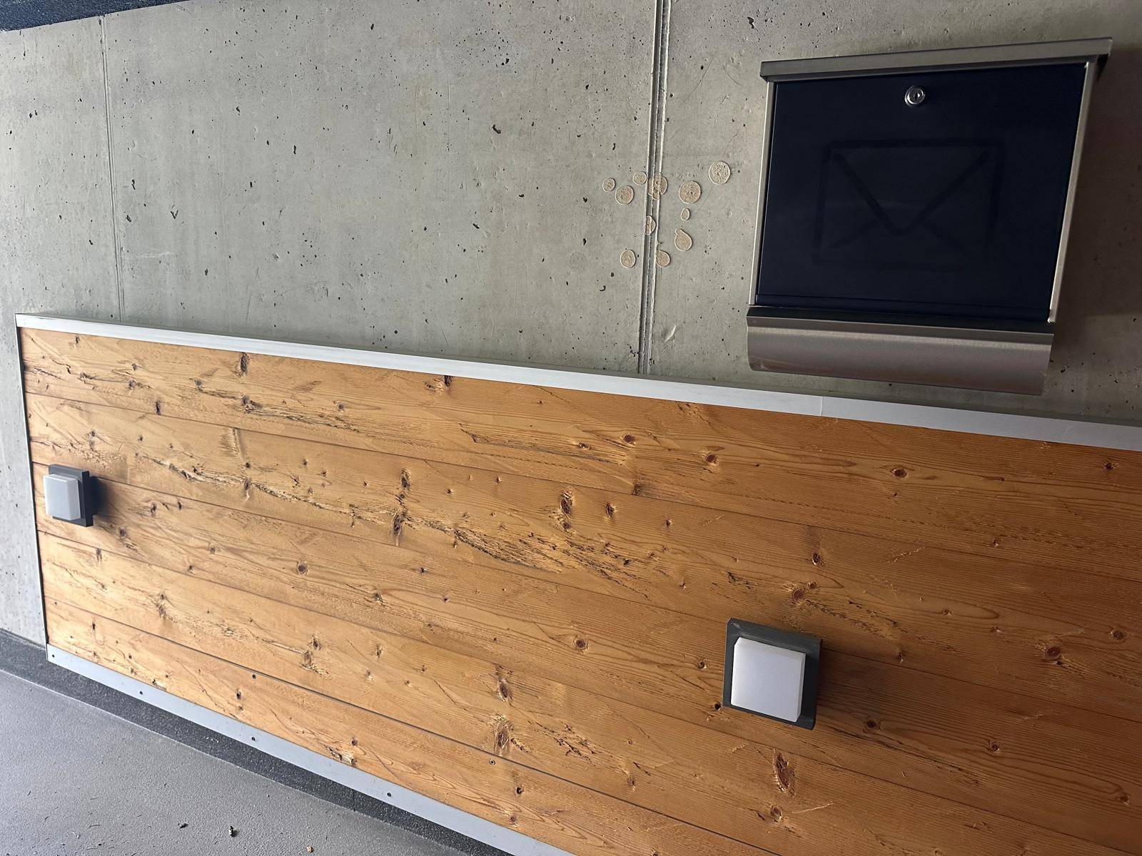 Außenwand mit Holzverkleidung, zwei Außenleuchten und einem modernen Briefkasten.