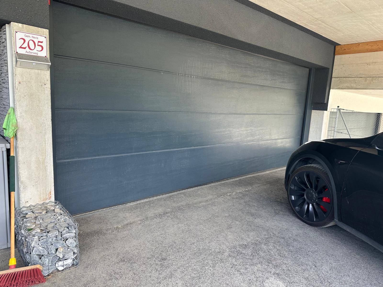 Garageneinfahrt mit dunklem Garagentor, Hausnummer und einem teilweise sichtbaren Elektrofahrzeug.