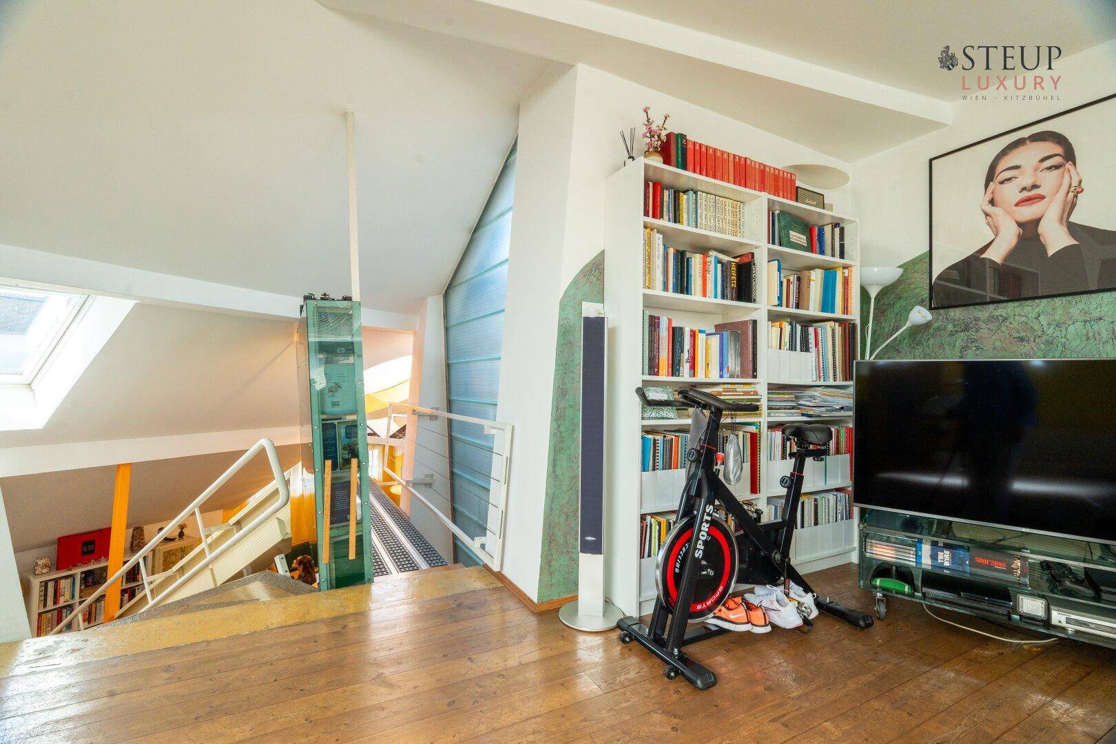 Offener Wohnbereich mit Bücherregal, Fernseher und Trainingsgerät, verbunden mit dem Treppenhaus.