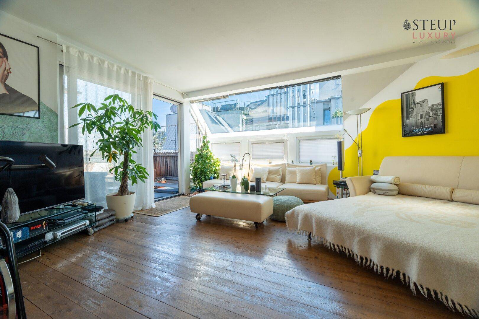 Helles Wohnzimmer mit Sofa, Fernseher und Zugang zu einem Außenbereich durch große Fenster.