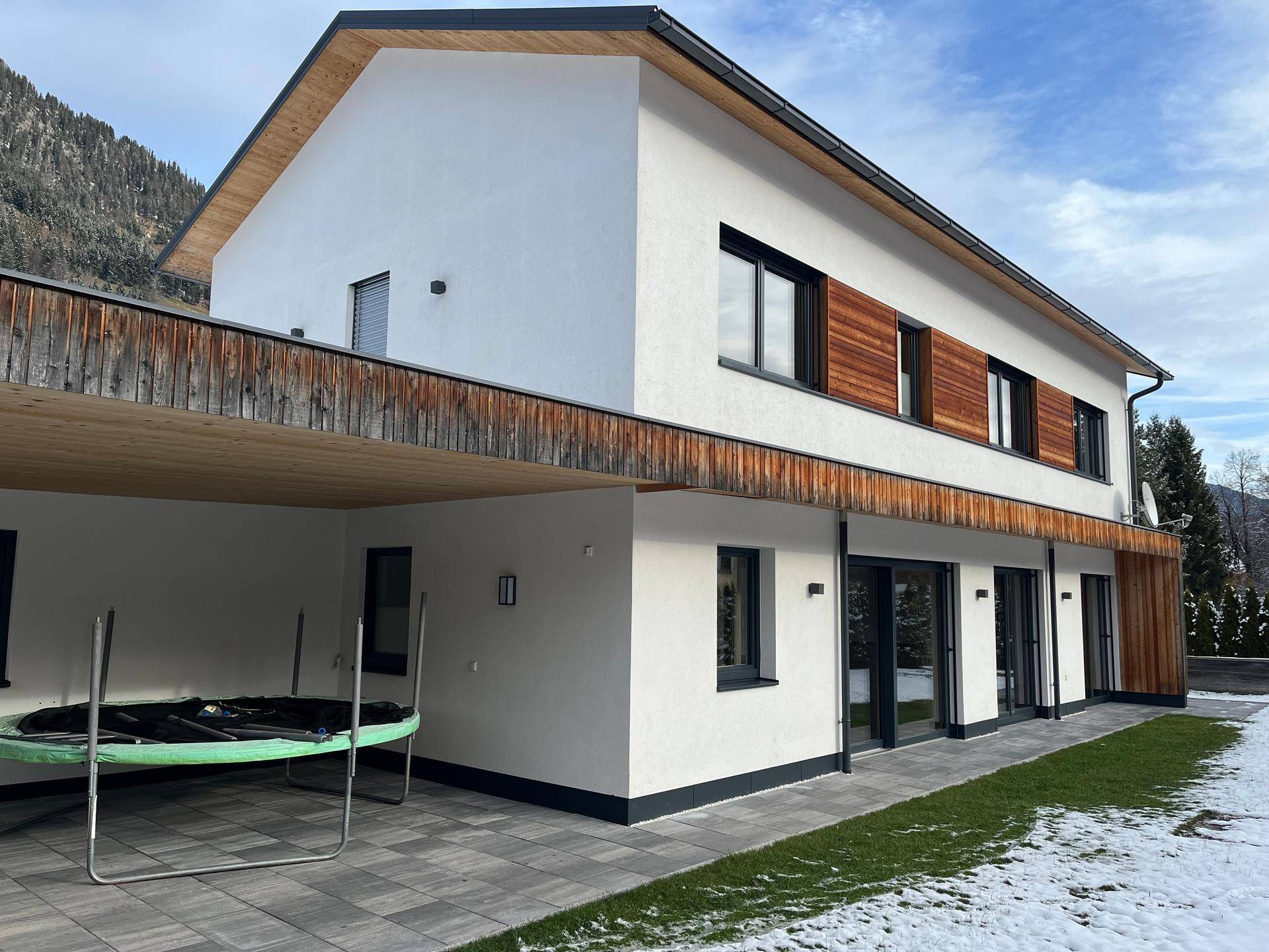 Modernes Einfamilienhaus mit weißer Fassade, Holzdetails und Carport in bergiger Umgebung.