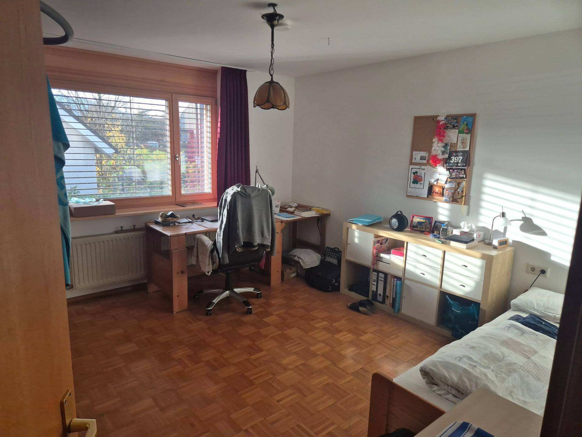 Helles Schlafzimmer mit Parkettboden, Schreibtisch und großem Fenster mit Blick ins Grüne.