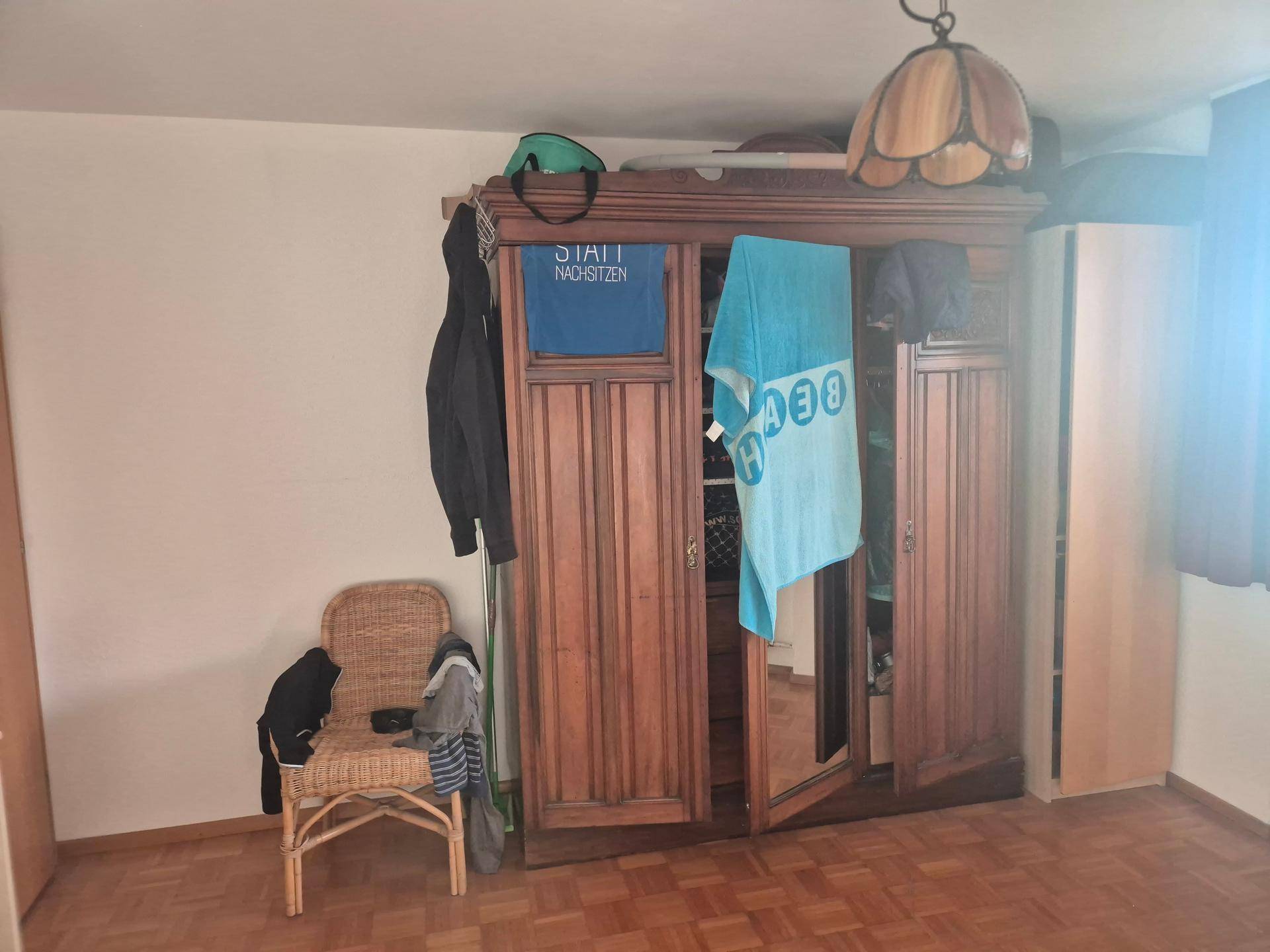 Schlafzimmer mit großem Holzschrank, Korbstuhl und klassischem Parkettboden.