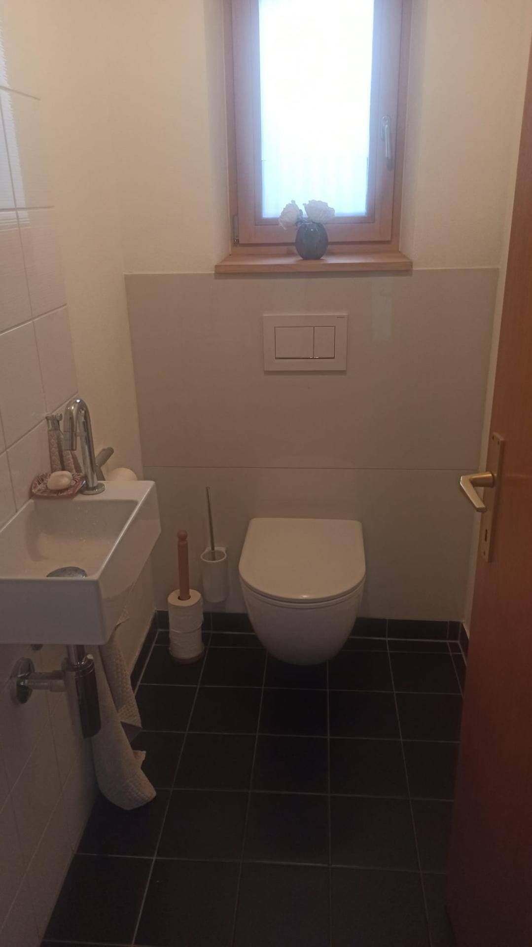 Separates WC mit wandhängendem WC, kleinem Waschbecken und Fenster.
