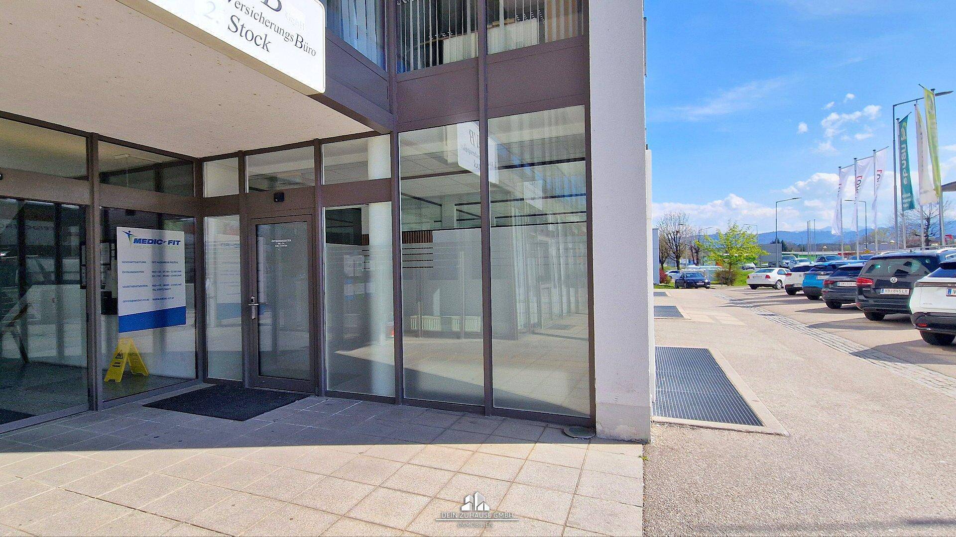Eingangsbereich eines Geschäftsgebäudes mit großen Glasfronten und einem gepflasterten Vorplatz.