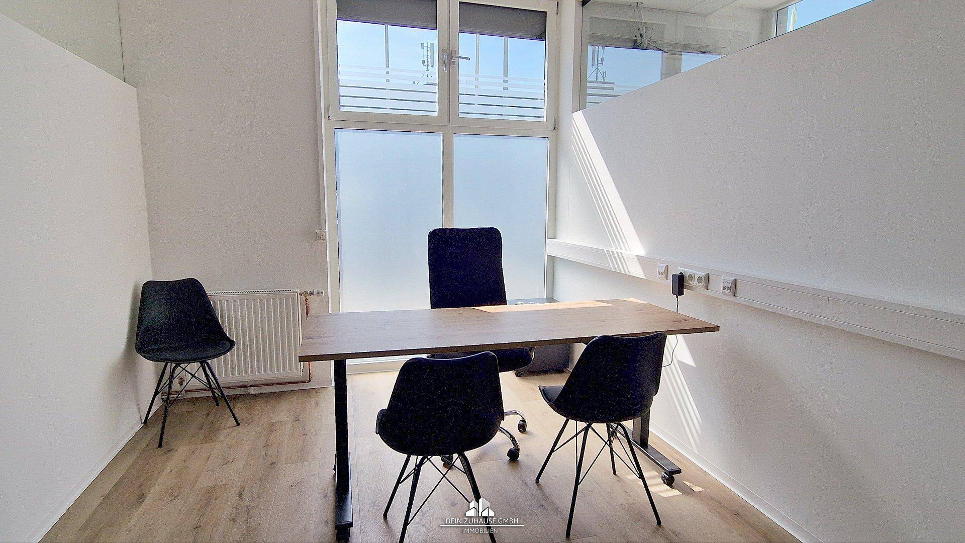Heller Büroraum mit einem Schreibtisch, drei Stühlen und großen Fenstern, die viel Tageslicht hereinlassen.