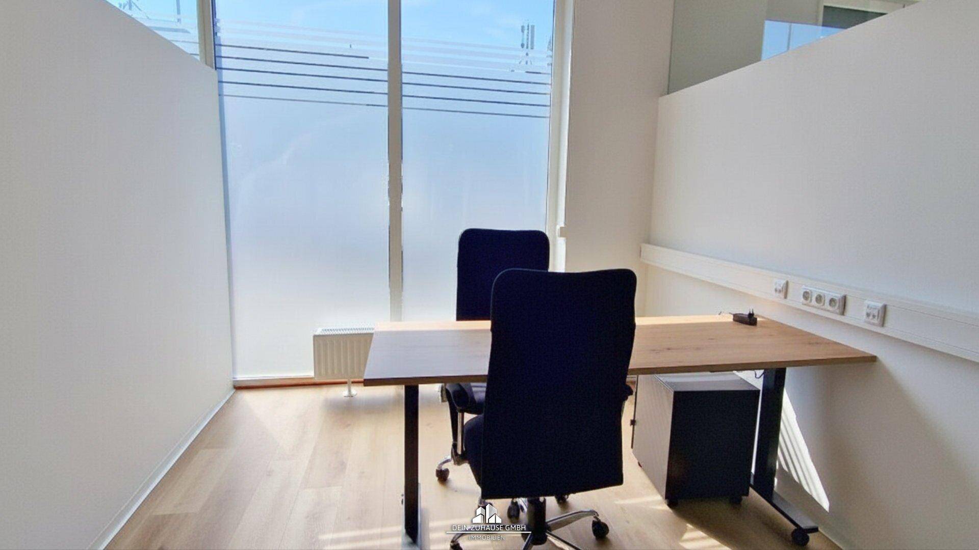 Moderner Büroraum mit einem Schreibtisch, zwei Bürostühlen und großen Fenstern, die für Helligkeit sorgen.