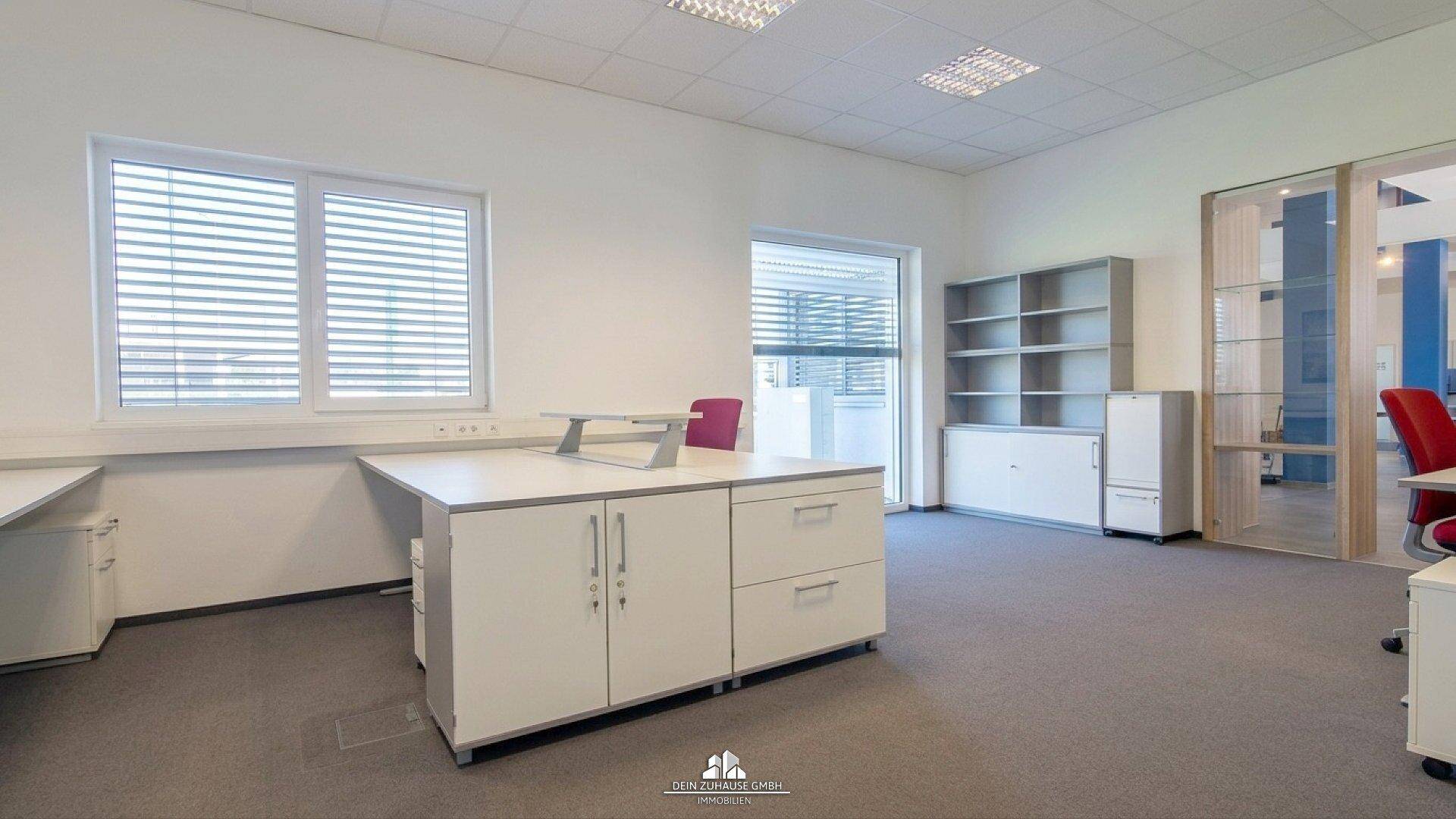 Geräumiges Büro mit Teppichboden, weißen Möbeln und Blick auf den Außenbereich durch große Fenster.