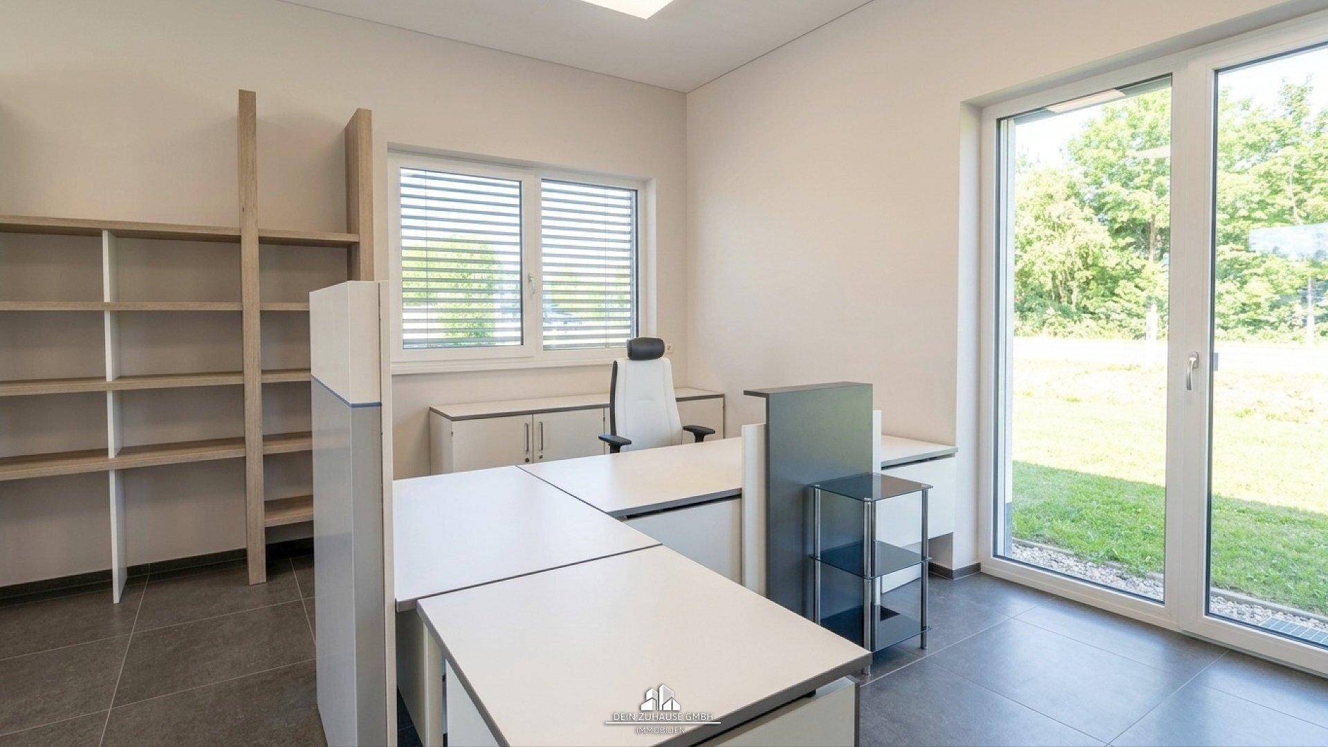 Modernes Büro mit Schreibtischen, Regalen und großen Fenstern mit Blick ins Grüne.