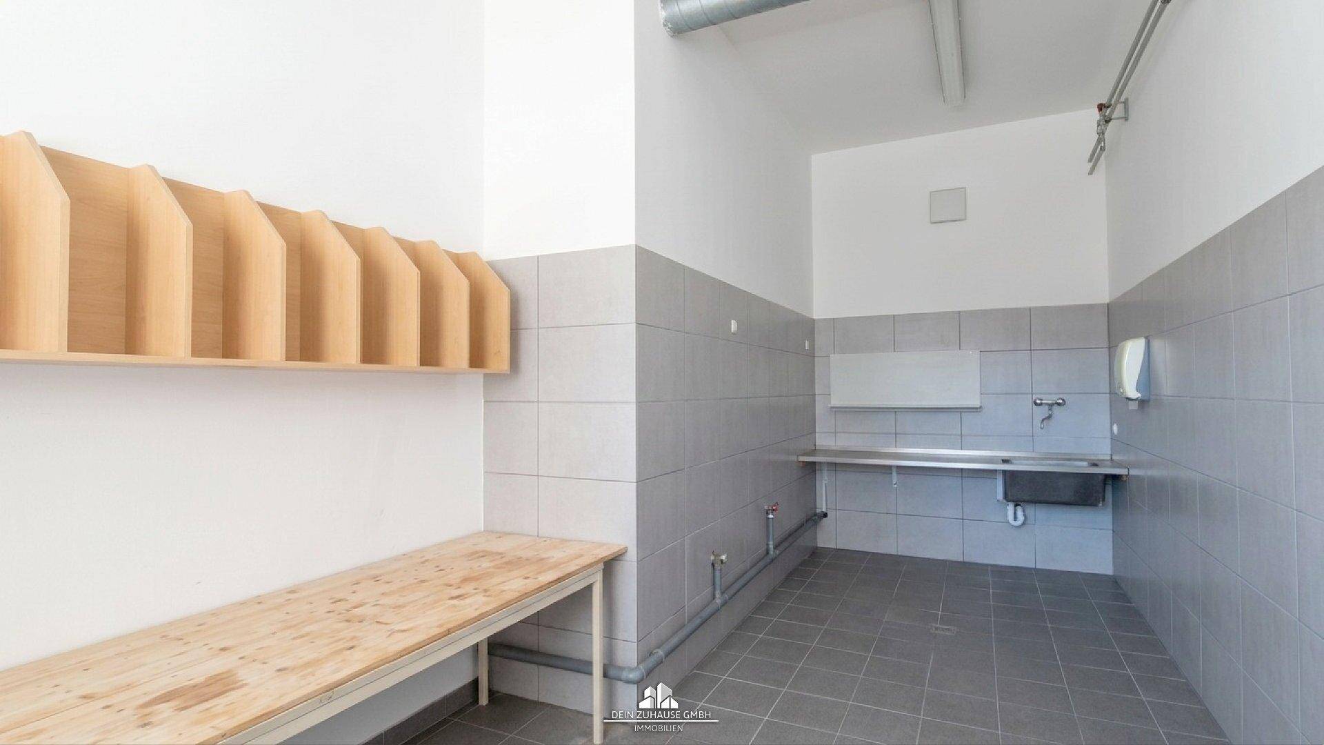Umkleideraum mit Holzbänken, Ablagefächern und einem Waschbeckenbereich.