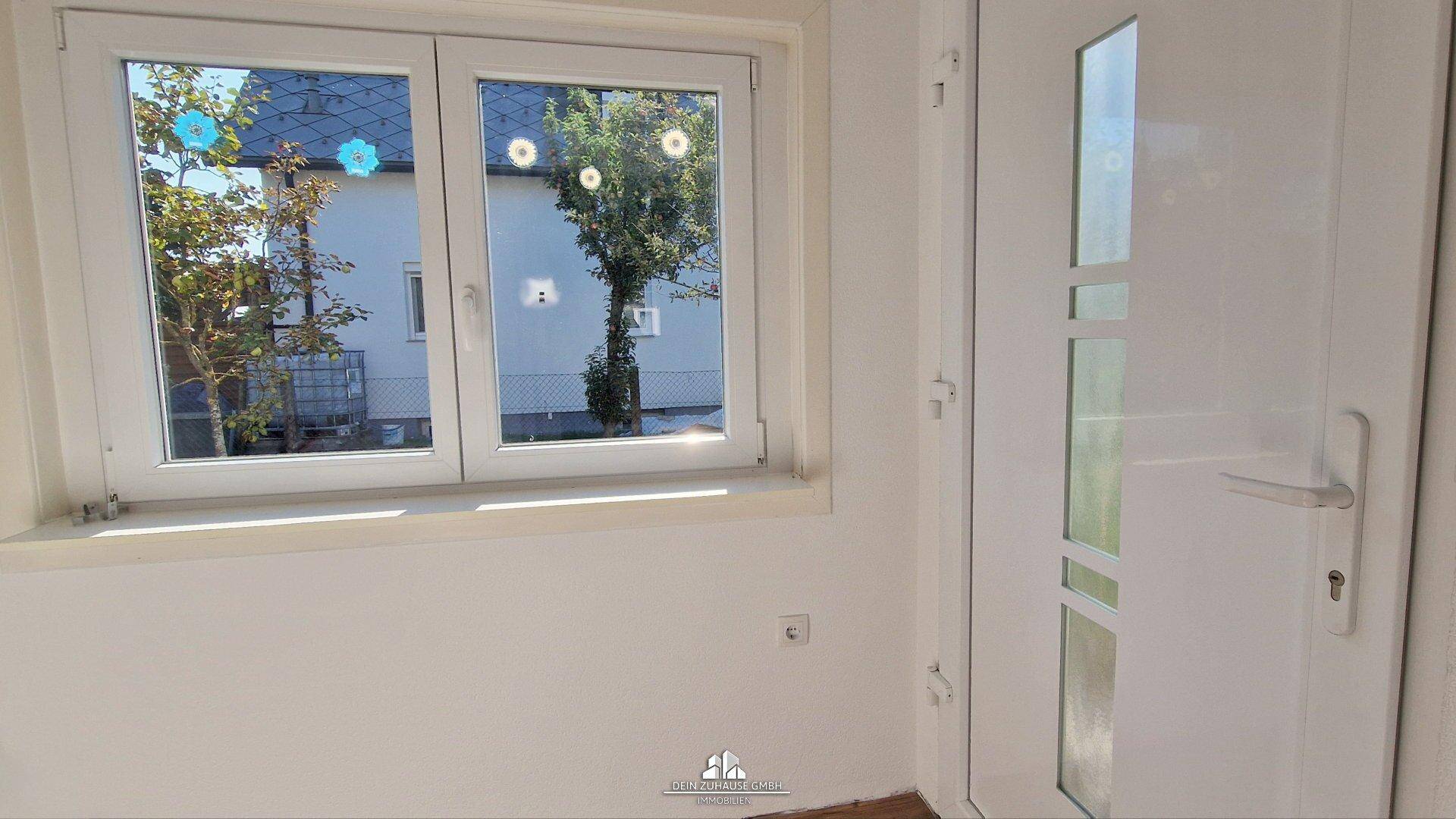 Heller Eingangsbereich mit weißer Tür und Fenster, das den Blick auf den Außenbereich freigibt.